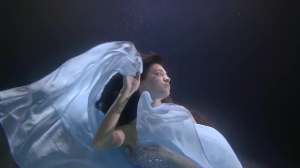 krásná dívka s dlouhými splývajícími vlasy a flitrovanými šaty se vznáší pod vodou. mává rukama a modrou látkou jako křídly