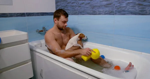 An adult dark-haired man with a beard, he is in the bathroom, sitting in a bath with foam and washes there with the dog, wipes the dog with a sponge, as well as two balls floating in the water.