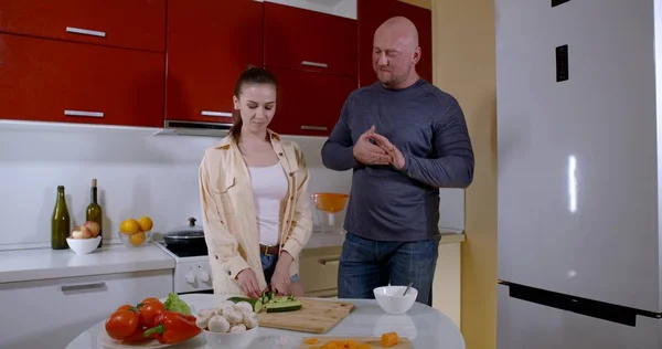 the couple is dressed in casual clothes and stands at the kitchen table. the girl cuts a cucumber, tries it. the man says something and takes a tomato