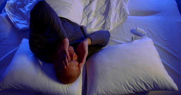 Close-up portrait of a bald, middle-aged man lying in bed with white linens and two pillows. He tosses and turns, holding his head. He has a headache and takes a pill.