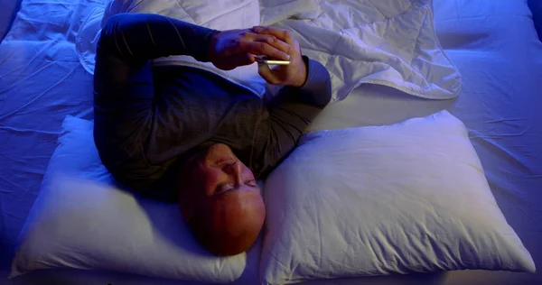 Close-up portrait of a bald, middle-aged man lying in bed with white linens and two pillows. He looks at his smartphone and is indignant waving his hand.
