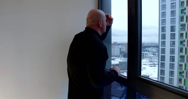 A bald middle-aged man is on a spacious balcony with large Windows, looking out the window leaning on it, outside the window is a new building. Hes wearing a black jacket.