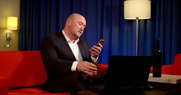 A bald middle-aged man is sitting in a room on a red sofa, wearing a business suit, a table in front of him, a laptop on it, and a bottle of alcohol. Hes on the phone, smiling, drinking from a glass.