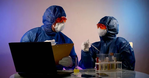 two people in protective clothing, respirators, glasses and gloves are sitting at a table. they discuss actions, one dripping from a pipette into a Petri dish, the other writing on a paper tablet
