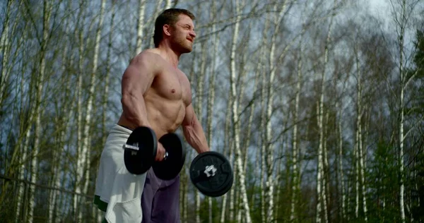 The man is on the street against the background of a forest of birches, he is wearing sweatpants, and on top of the naked torso, he makes swings with his hands holding two large dumbbells.