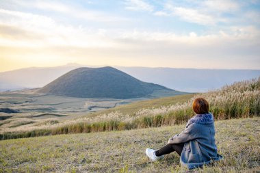 Aso Nakadake Dağı, Kumamoto, Kyushu, Japonya 'daki dumanlı aktif volkan arka planının yanında oturan sevimli Japon kız.)