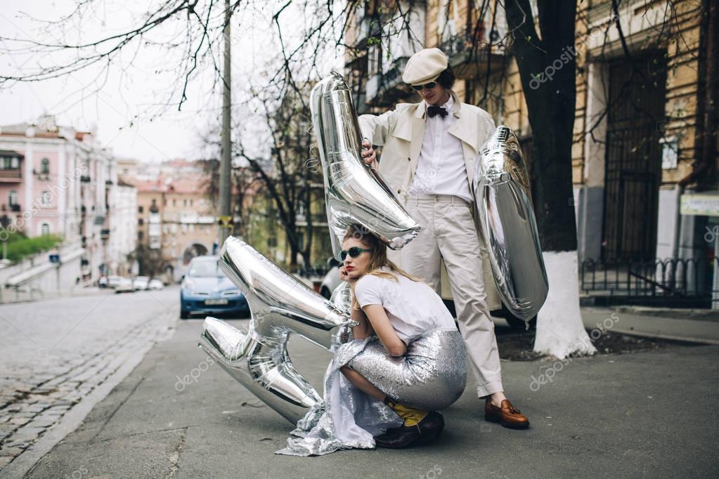 Stylish woman crouching with standing man and silver balloons on city street