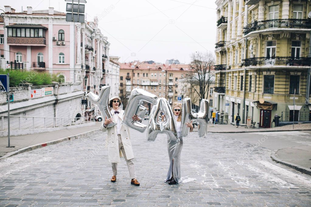 Stylish couple posing with silver balloons in shape of letters on city street