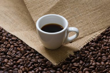 Kahve çekirdekleri ve kaba bir çuval bezi üzerine beyaz bir fincan kahve. Kahve bileşimi.