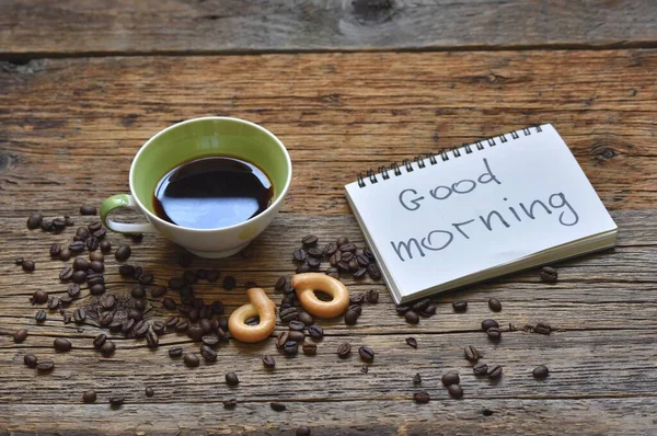 A fresh Cup of brewed coffee on a wooden background and a greeting note in the notebook Good Morning.