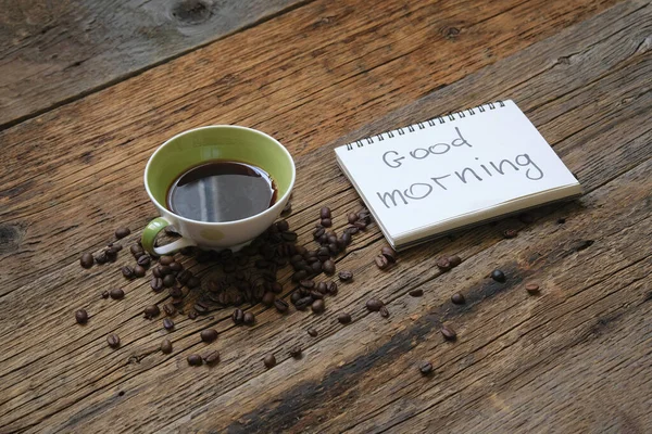 A fresh Cup of brewed coffee on a wooden background and a greeting note in the notebook Good Morning.