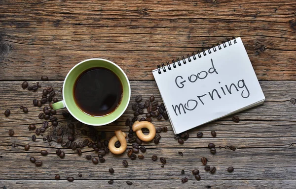 A fresh Cup of brewed coffee on a wooden background and a greeting note in the notebook Good Morning.