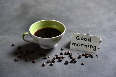 Koyu arkaplanda bir fincan taze kahve ve defterde bir karşılama notu. Günaydın. 