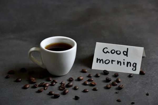 A fresh Cup of brewed coffee on a dark background and a greeting note in the notebook Good Morning