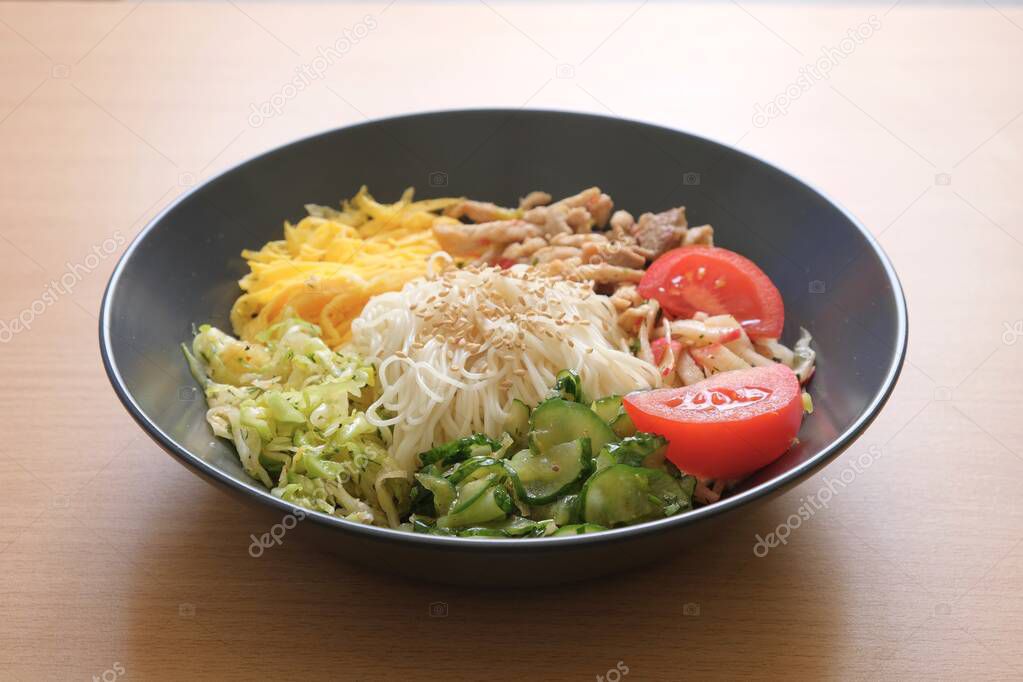 Traditional Korean dish Kuksi on a wooden background (one of the types of Kuksi). Ingredients-pasta, vegetable salads of cucumbers, cabbage, radish, meat, omelet. The broth is served separately.