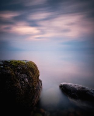 Ön planda yosun olan taşlar ve arka planda denizin üzerinde esen bulutlar.