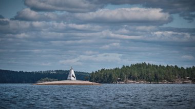Arka planda orman olan gölün üzerinde küçük beyaz bir deniz feneri. Gökyüzünde beyaz bulutlar