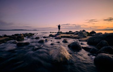 Bir kişi durur ve deniz kenarında denize bakar. Arka planda günbatımı, taşlarda ve önde bir nehir vardır.