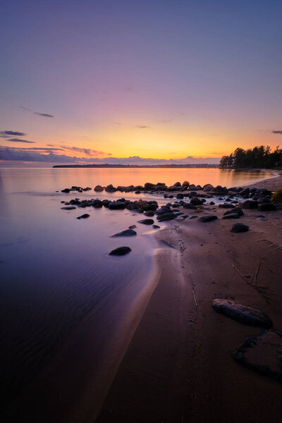 Sunset at a lake with sandy beach with large stones leading through the picture to a forest