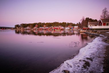 Gün batımında fotoğraf ön planda buzla kaplı bir körfezin üzerinde ve körfezin kenarında küçük kırmızı kulübeler.