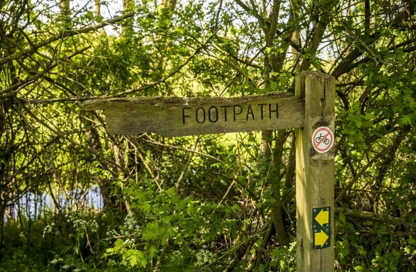 A wooden footpath sign in a shady wooded area with way markers and a no cycling sign on the post