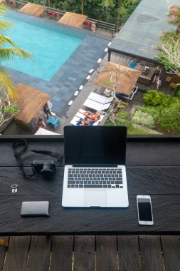 Yüzme havuzu arka planında siyah ahşap masada kameralı ve akıllı telefonlu bir laptop aç