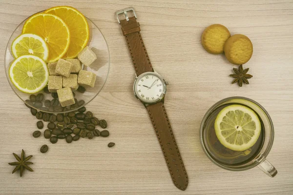 Flat lay. Men\'s watches with leather strap. Cup of tea with lemon. Slices of lemon and orange on a saucer. Coffee beans, cookies and pieces of sugar on the table