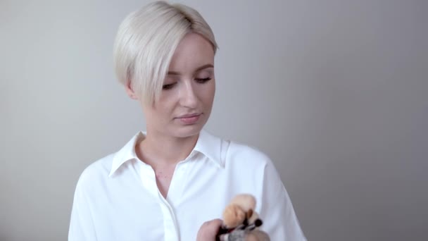 Menina loira de shorthaired bonito olha para sua coleção de escovas essenciais para a aplicação de maquiagem diária . — Vídeo de Stock