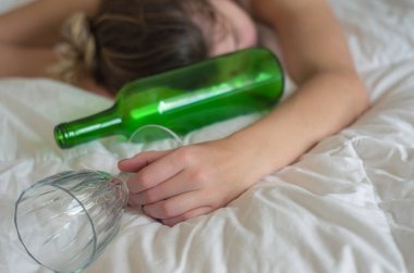 Great concept of alcohol abuse. Young woman, blond hair, fainted in bed after drinking too much alcohol. Glass of wine in hand, bottle of wine. clipart