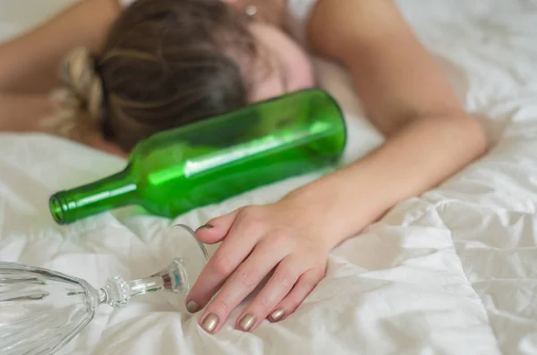 Great concept of alcohol abuse. Young woman, blond hair, fainted in bed after drinking too much alcohol. Glass of wine in hand, bottle of wine.