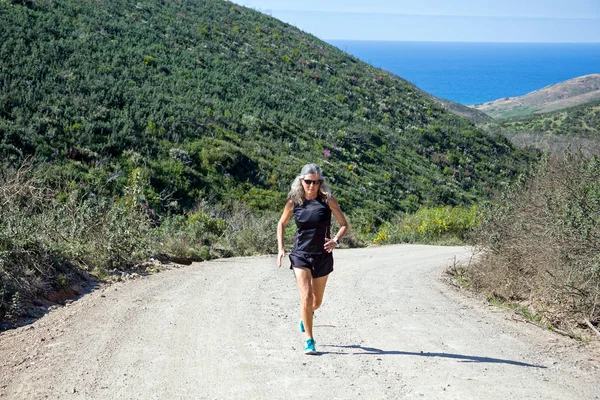 Sportive mature woman jogging uphill at the atlantic ocean — Stock Photo, Image
