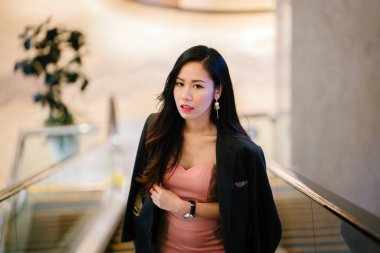 şık ve modaya genç Çinli Asyalı kadın bir yürüyen merdiven üzerinde. Omuzları üzerinde bol dökümlü ve gülümseyen bir adamın ceketi ile güzel bir pembe elbise giyiyor.