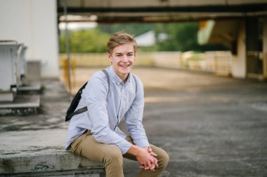 dikey bir genç ve çekici beyaz beyaz adam (turist veya öğrenci) oturma ve gündoğumu veya günbatımı Asya beton bir araba parkta gülüp. O gülümsüyor ve rahat.