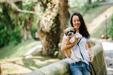 Pet shih tzuh köpeğini gün yeşilliklerle dolu bir parkta ve bitkiler içinde belgili tanımlık geçmiş olan bir genç, çekici ve güzel pan Asya kadın portresi.