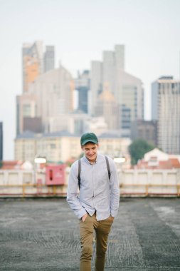 dikey bir genç ve çekici beyaz beyaz adam (turist veya öğrenci) oturma ve gündoğumu veya günbatımı Asya beton bir araba parkta gülüp. O gülümsüyor ve rahat.