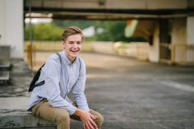 dikey bir genç ve çekici beyaz beyaz adam (turist veya öğrenci) oturma ve gündoğumu veya günbatımı Asya beton bir araba parkta gülüp. O gülümsüyor ve rahat.