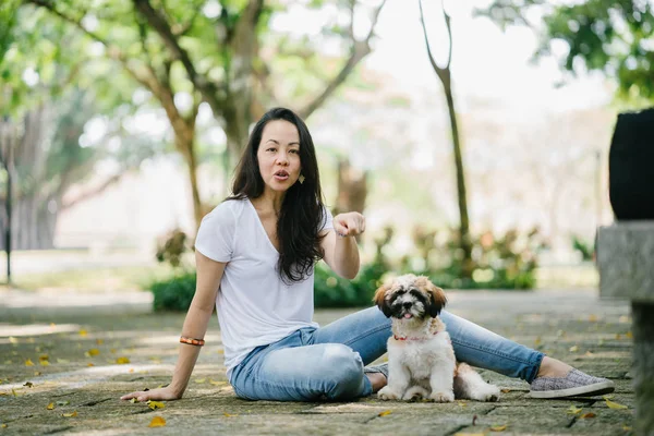 Portrait of a young Pan Asian woman sitting with her young shih tzuh puppy dog in the park in the day. She is smiling and they are both enjoying the warm day.