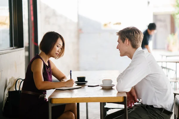 An Asian woman is interviewing a young Caucasian white man for an internship job in Asia. They are sitting at a desk in the shade outside in the day and talking over coffee.
