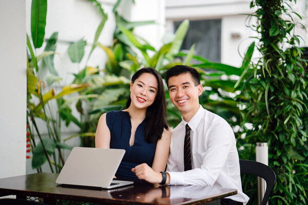 Portrait of two smiling business people (lawyers, consultants, etc) or couple smiling mid discussion over a laptop in an office with greenery in the background. They are focused on the laptop.