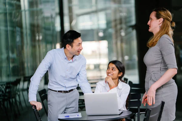 3 people from a diverse team are having a discussion around a laptop computer in the day in an open area. They are energetic and cheerful as they smile and talk.
