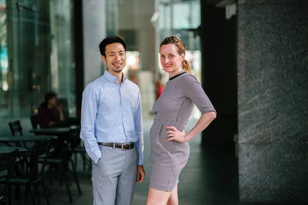 Portrait of two coworkers, team mates or colleagues standing in their office during day. One is an woman, and the other a Chinese man (diverse). They are smiling in a relaxed way.