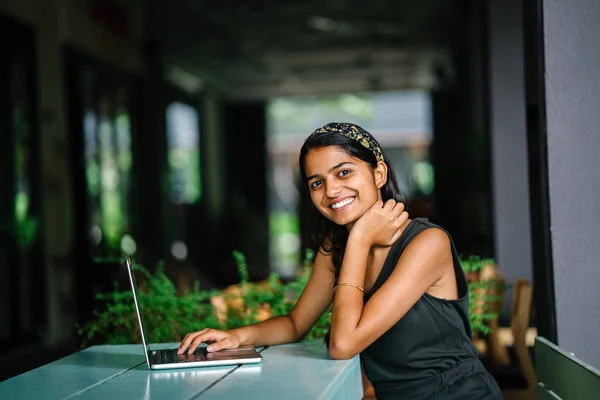 Candid portrait of an attractive and young Indian Asian professional woman work on her laptop in a trendy cafe or coworking space with teal wooden furniture. She is smiling and looking away.
