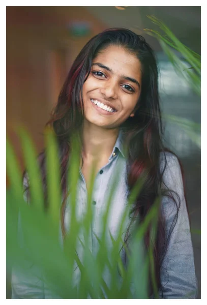 バック グラウンドでの植物の近くの若いインド人女性 彼女は若い 魅力的で率直な自然な方法で笑顔です 彼女は青いシャツを着ています — ストック写真