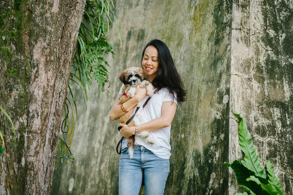 Portrait of a young Pan Asian woman sitting with her young shih tzuh puppy dog in the park in the day. She is smiling and they are both enjoying the warm day.