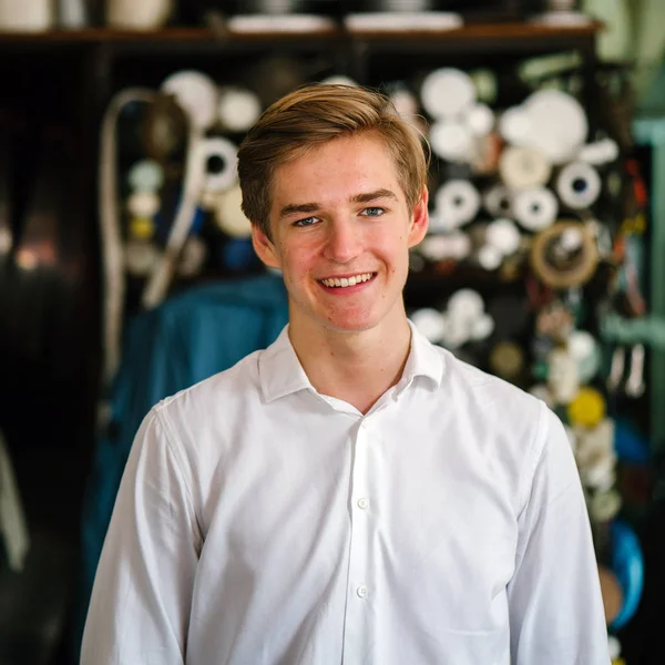 young Caucasian man stands in front of a metalworking workshop with metal pipes in the background. He is smiling as he looks into the camera. He is young and photogenic.