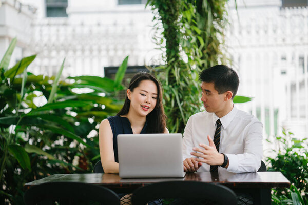 Portrait of two smiling business people (lawyers, consultants, etc) or couple smiling mid discussion over a laptop in an office with greenery in the background. They are focused on the laptop.