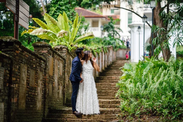 interracial couple (Indian man, Chinese woman) pose for wedding photo in a park in the day. They are on old stone steps and and smiling and laughing as they take photos.