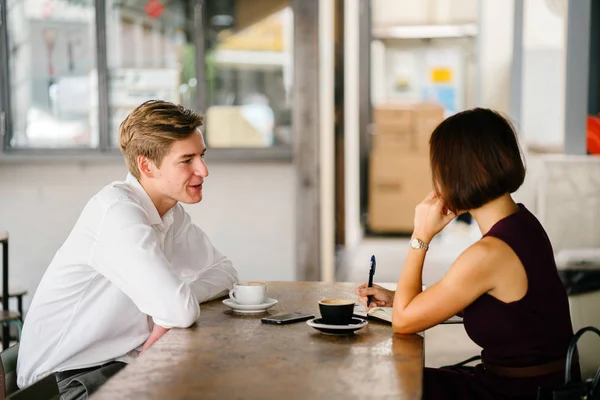 An Asian woman is interviewing a young Caucasian white man for an internship job in Asia. They are sitting at a desk in the shade outside in the day and talking over coffee.