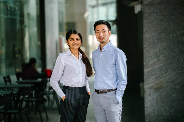 Portrait of two coworkers, team mates or colleagues standing in their office during day. One is an Indian woman, and the other a Chinese man (diverse). They are smiling in a relaxed way.