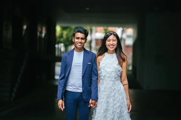 portrait of a young interracial couple getting married. An Indian man and his Chinese wife pose for a photograph in the day along the street against a dark background.